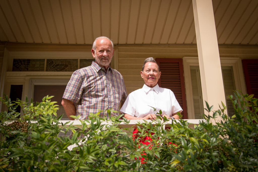 A light-skinned couple stands on the front porch of their house and overlook a green hedge of plants.