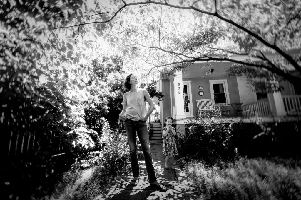 A tall thin, light-skinned woman stands in her front yard as her toddler looks on.