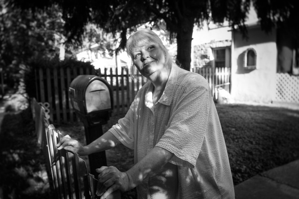 A light-skinned older woman with gray hair stands at her mailbox and looks up at the sky.