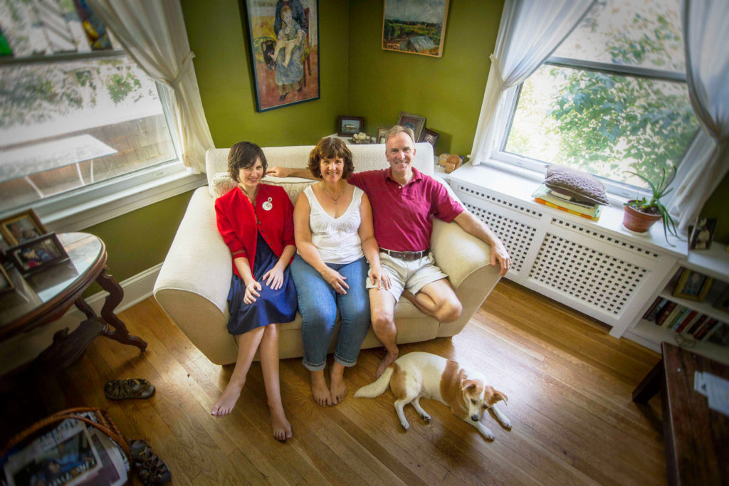 Two women and a man look up from an oversized chair in a house as a dog rests at their feet.