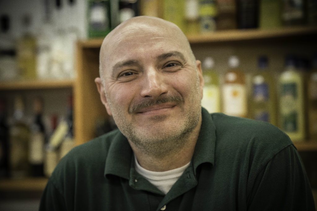 A fair-skinned man with a bald head and green, collared shirt smiles as he sits in front of a shelf with wine bottles. 