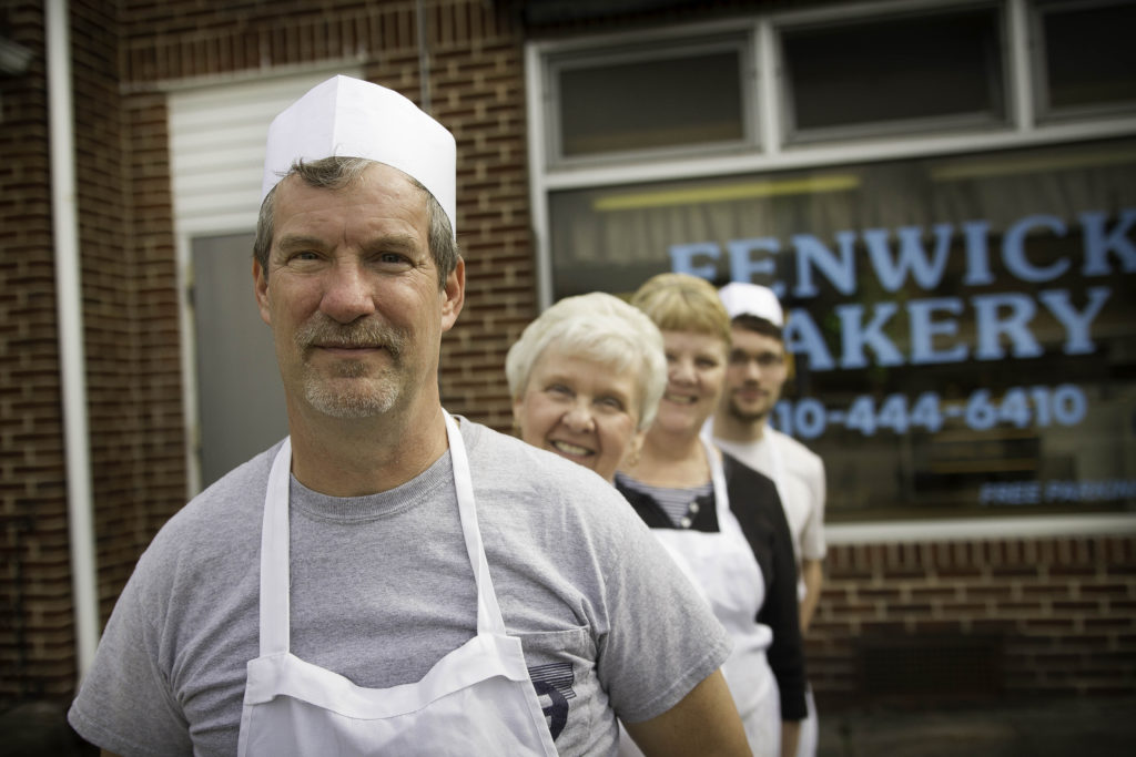 Four fair-skinned people stand in a row outside of a bakery. The man in front wears a white chef's hat and white apron.