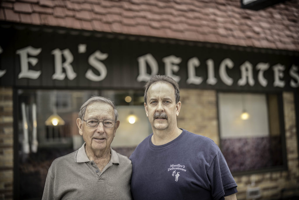 Two fair-skinned men, one older and one younger, stand in front of a sign that reads Mueller's Delicatessen. 