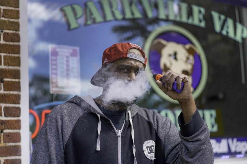 A brown-skinned man with a red baseball cap and gray, zip-up sweatshirt stands in a puff of smoke.